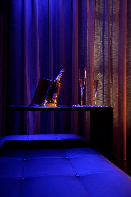 Duas flutes e garrafa de champanhe sobre uma mesa de apoio vistos da cama, iluminada com luzes roxas, na suite Nova Iorque.