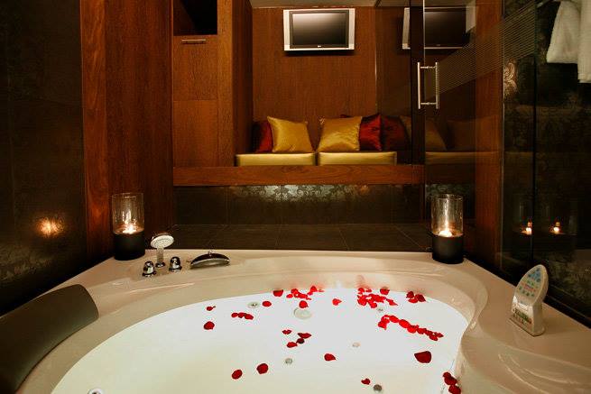 Jacuzzi iluminado e decorado com pétalas de rosa vermelha e um banco almofadado ao fundo, na suite Veneza