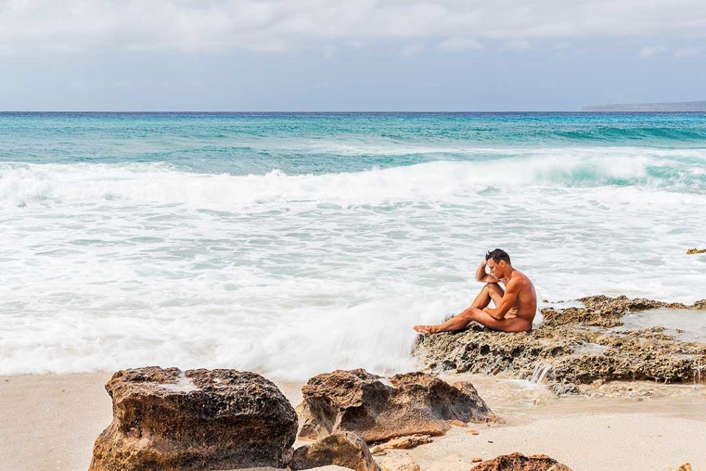 Homem nu sentado numa rocha à beira-mar